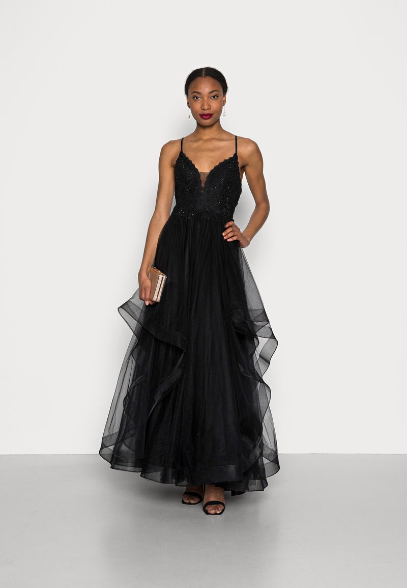 Abito da sera nero con scollo profondo, corpetto decorato e gonna a strati in tulle. La modella tiene una pochette metallica e indossa tacchi neri.