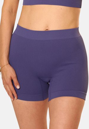 Frau trägt gerippte, lila, hoch taillierte Biker-Shorts und passendes Crop-Top, Hand ruht auf dem Oberschenkel, vor einfarbigem Hintergrund.