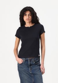 Tamsiai mėlynas trumpas rankovėmis viršus su apvaliu iškirpimu, su bangomis apkarpytais kraštais ir mažu logotipo žymekliu ant apačios. Medžiaga atrodo lygi ir prigludusi.