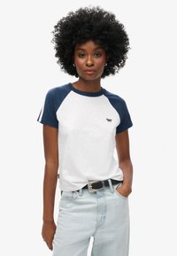 Hvid t-shirt med marineblå raglanærmer, rund hals og lille logo. Bæres med højtaljede lyseblå jeans og et sort bælte.