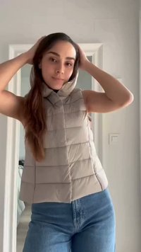 Femme ajuste la capuche d'une veste sans manches beige matelassée, porte un jean bleu, debout à l'intérieur près d'une porte blanche et d'un interrupteur.