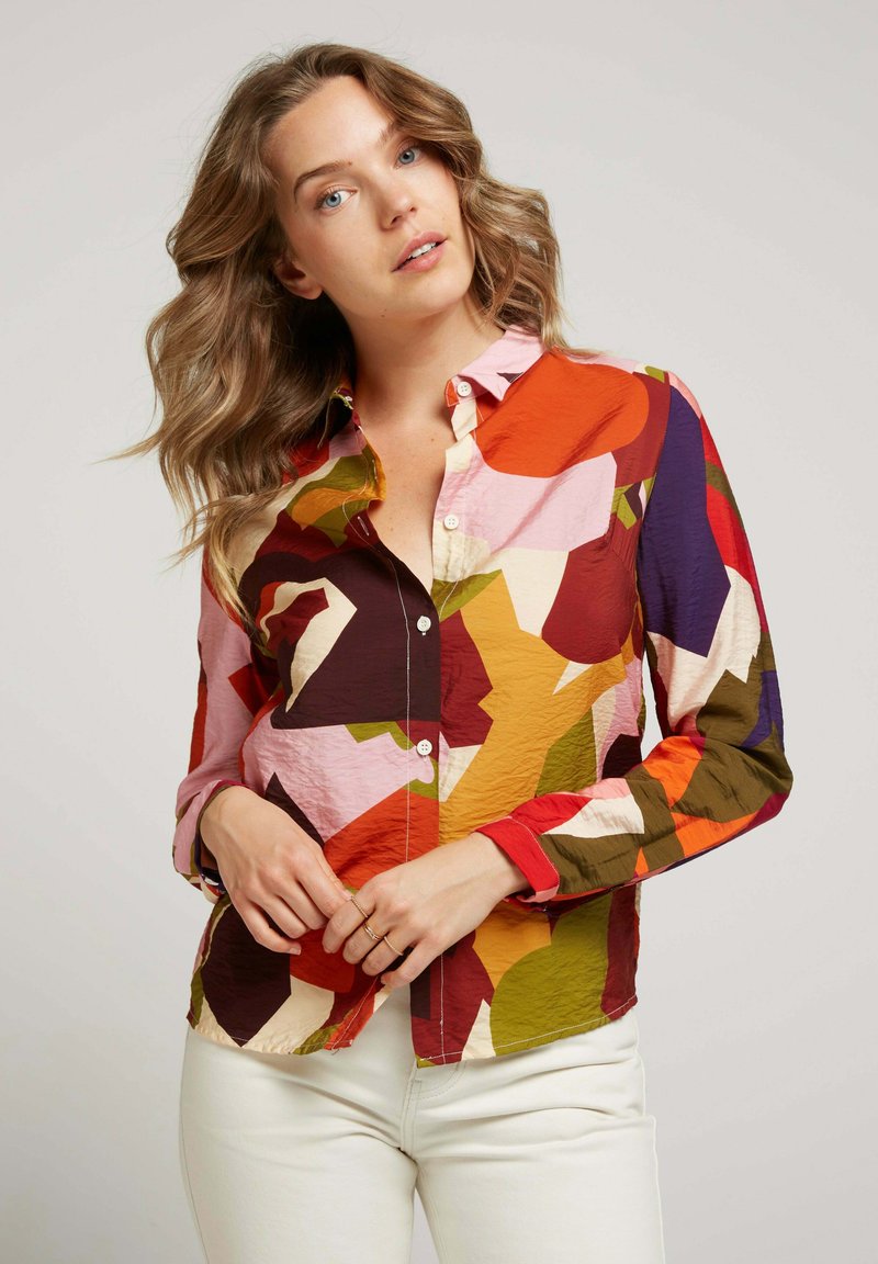 Chemise colorée à manches longues présentant un mélange de motifs abstraits en rouge, orange, rose et vert, avec un col et des boutons.