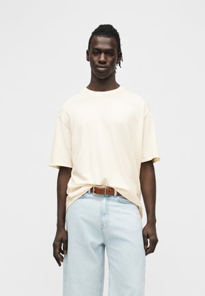 Giovane uomo con pelle scura e dreadlocks corti indossa una maglietta beige larga infilata in jeans chiari con cintura marrone, in piedi davanti a uno sfondo bianco.