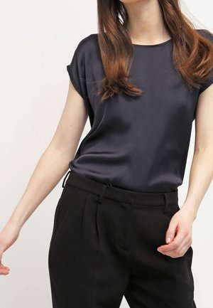 Personne portant un chemisier en satin noir à manches courtes rentré dans un pantalon noir taille haute et ajusté, sur un fond uni.