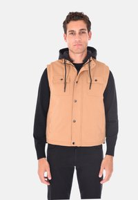 Gilet beige con superficie testurizzata, due tasche frontali, cappuccio nero e cordini, indossato su una maglietta nera a maniche lunghe.