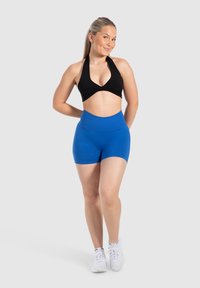 Zwarte haltercrop top en hooggetailleerde blauwe shorts, beide in een gladde stof, gecombineerd met witte sneakers op een neutrale achtergrond.