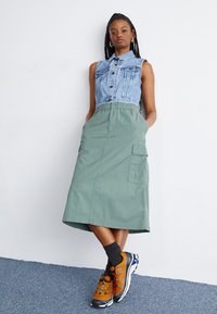 Jachetă din denim fără mâneci, cu un design scurt, asortată cu o fustă cargo verde, până la genunchi, cu buzunare. Pantofi sport portocalii și negri completează ținuta.