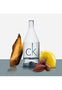 Bouteille en verre clair avec un bouchon bleu et une étiquette 'ck'; à proximité, un morceau de bois, un citron jaune et des cabosses de cacao brunes sur une surface réfléchissante.
