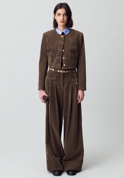 Giacca corta in corduroy marrone con bottoni dorati e due tasche frontali, abbinata a pantaloni a gamba larga coordinati e una cintura a catena, su uno sfondo neutro.
