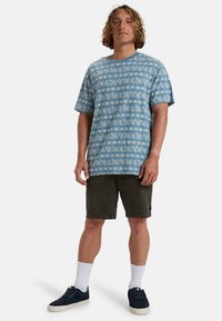 Blå kortermet skjorte med blad- og diamantmønster, laget av bomull. Kombinert med mørke shorts og hvite sokker, stående på en ensfarget bakgrunn.