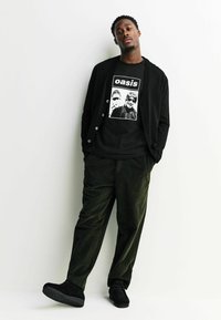 Cardigan negru peste un tricou negru cu un imprimeu grafic, asortat cu pantaloni de catifea verde închis și încălțăminte neagră.