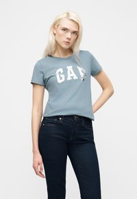 Ξανθιά γυναίκα που φοράει μπλε μπλουζάκι GAP και σκούρο μπλε τζιν, στέκεται με το ένα χέρι στη μέση μπροστά σε απλό λευκό φόντο.