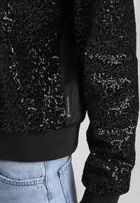 Schwarzes Sweatshirt mit Pailletten und glatter Textur, gerippten Bündchen und Saum. Mit Logo-Label an der Seite und lässiger Passform. Kombiniert mit hellblauen Jeans.