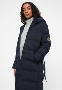 Mørkeblå puffjakke med hætte, lynlås-lukning og ribbede manchetter. Har et logo-patch på venstre arm og en quilte-design.