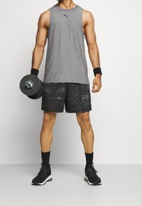 Šedé bezramínkové tílko s černým logem, černé kraťasy s texturovaným vzorem a černé sportovní boty. Drží černou činku.