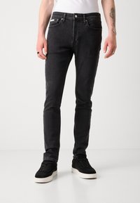 Vaqueros negros de corte slim fabricados en denim con una textura suave, que cuentan con bolsillos estándar y un botón de metal para el cierre. Combinados con zapatillas negras.