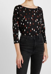 Schwarzes Langarmoberteil mit Rundhalsausschnitt, das mit einem gepunkteten Muster in Rot, Weiß und Beige versehen ist. Kombiniert mit hochgeschnittenen schwarzen Jeans.