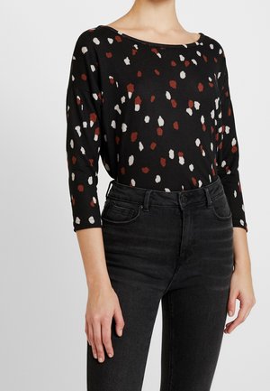 Schwarzes Langarmoberteil mit Rundhalsausschnitt, das mit einem gepunkteten Muster in Rot, Weiß und Beige versehen ist. Kombiniert mit hochgeschnittenen schwarzen Jeans.