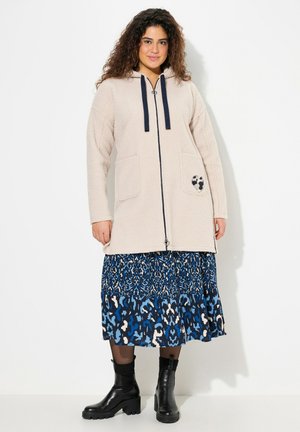 Beige fleece vetoketjullinen takki hupulla, laivaston siniset nauhat, kaksi etutaskua, yhdistetty siniseen kuvioituun hameeseen ja mustiin nilkkureihin.
