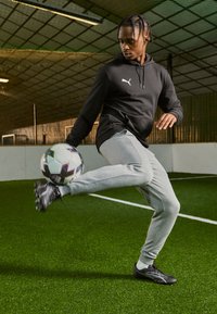 Melnā kapucēta džemperis, pelēkas sporta bikses un melni futbols tēvu apavi, ko valkā persona, kas sit futbolu zaļā mākslīgā zālē iekštelpās.