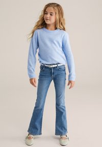 Junge Mädchen steht in einem hellblauen Pullover, blauen Jeans mit weißem Gürtel und blassen Schuhen, schaut zur Seite vor einem beigen Hintergrund.
