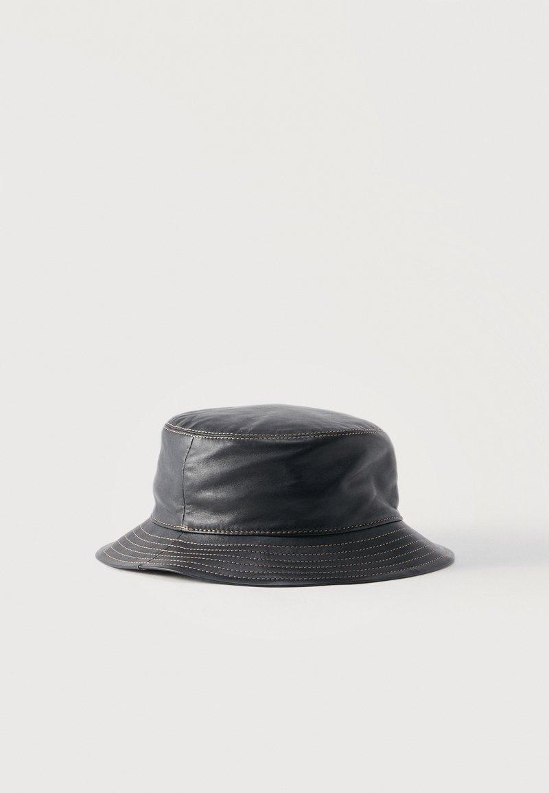Cappello a secchiello in pelle nera con cuciture marroni visibili sulla parte superiore e intorno alla tesa, posato su una superficie chiara e semplice.