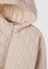 Giacca beige trapuntata con cappuccio, caratterizzata da un motivo a diamante e piccoli dettagli in argento. Chiusura con zip e fodera interna liscia.