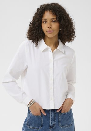 Woman with curly hair wearing a white button-up shirt and blue jeans, standing with hands in front pockets against a white background.