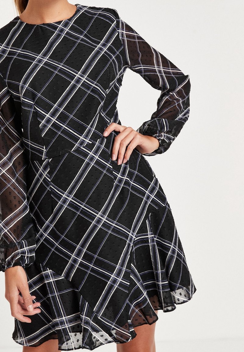 Robe à carreaux noirs avec des manches longues transparentes, des pois texturés, un ourlet asymétrique et des lignes blanches et violettes dans un motif à carreaux.