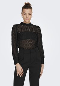 Svart topp med teksturert mønster, lange ermer, høy hals og semi-transparent design, kombinert med høy midje svarte bukser.