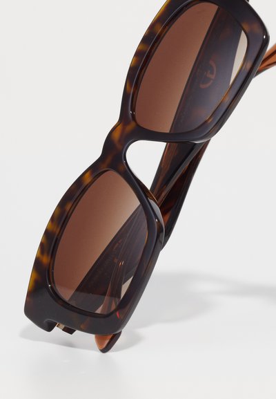 Lunettes de soleil rectangulaires en écaille de tortue avec des verres teintés marron, pliées avec une branche derrière la monture sur une surface blanche.