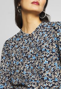 Kwiatowa bluzka w granatowym kolorze z niebieskimi i beżowymi wzorami. Posiada wysoki kołnierz, guziki oraz marszczone rękawy dla dodatkowej faktury.