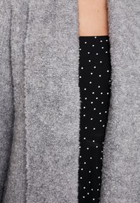 Mjukt, texturerat grått cardigan med öppen front, som visar en svart klänning med vita prickar underneath. Inga knappar eller hårdvara synliga.
