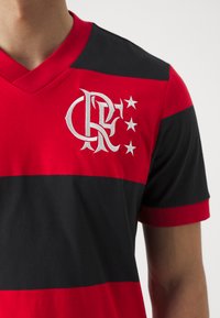 Röd och svart randig t-shirt med kort ärm och v-ringning; har en broderad logotyp och stjärnor i vitt på vänster bröst.