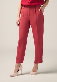 Pantaloni su misura rossi con vestibilità dritta, dotati di tasche laterali e chiusura con bottone. Abbinati a tacchi alti rosa chiaro.