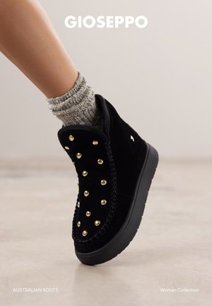 Botte de cheville en daim noir avec des clous dorés, chaussette en tricot texturé, bout arrondi et semelle épaisse noire, présentant des coutures détaillées le long des bords.