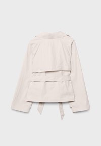 Veste femme beige clair à manches longues, rabat pare-tempête à l'arrière et ceinture à la taille suspendue, présentée à plat sur un fond clair.