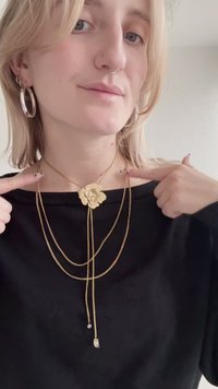 Χρυσό στρωματοποιημένο κολιέ με φυτικό μενταγιόν που έχει πολύτιμο λίθο στο κέντρο, φορεμένο πάνω από μαύρο τοπ, συμπληρωμένο με ασημένια σκουλαρίκια κρίκους.