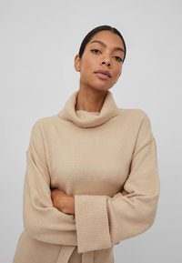 Pull en tricot beige avec un col roulé haut et des manches larges. Présente des accents côtelés aux poignets et une coupe détendue. Texture douce.