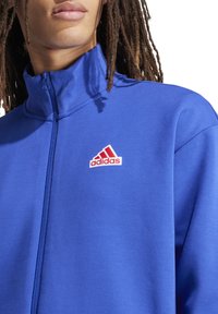 Blauwe ritsjack gemaakt van zacht materiaal. Kenmerkt zich door een hoge kraag en een rood Adidas-logo op de linkerkant van de borst. Soepele textuur en een strakke pasvorm.