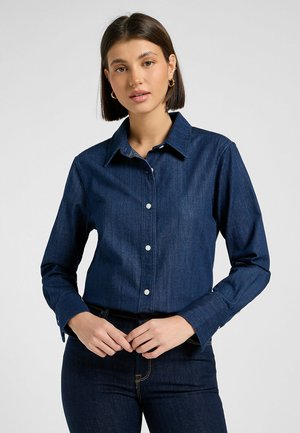 Camicia in denim blu scuro tagliata, con colletto, maniche lunghe e bottoni bianchi; texture liscia con dettagli di cucitura sottili.