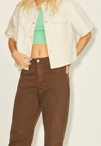 Light beige short-sleeve jacket with silver snap buttons and front pocket, paired with a green crop top and brown high-waisted pants.