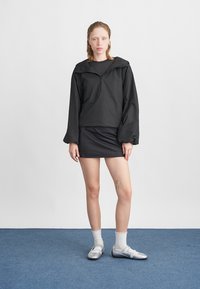 Czarny pulower z zamkiem błyskawicznym na ćwierć, z szerokimi rękawami, zestawiony z dopasowaną czarną spódnicą i błyszczącymi srebrnymi butami. Tło jest jednolite, białe.