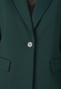 Blazer vert foncé avec un col cranté, une fermeture avec un seul bouton rond et deux poches frontales. Tissu lisse avec une coupe ajustée.