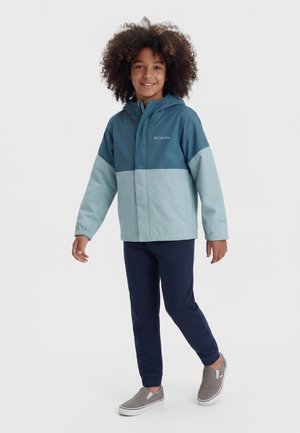 Dieťa s kučeravými vlasmi oblečené v modrej nepremokavej bunde s farebnými blokmi, tmavomodré teplákové nohavice a sivé slip-on topánky, stojí a usmieva sa.
