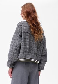 Kobieta z długimi, kręconymi czerwonymi włosami, nosząca okulary i szary marynarski żakiet w kratę z paskowym wykończeniem, stojąca na tle białym.