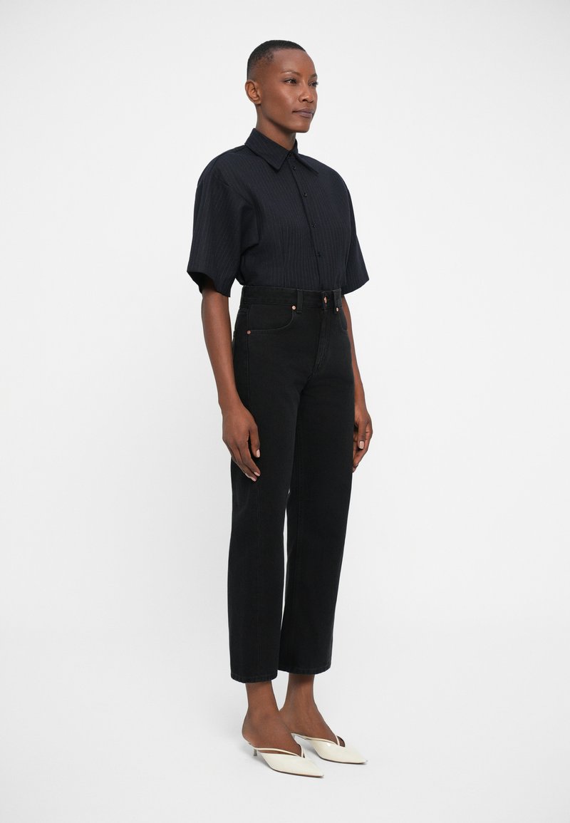 Chemise noire à manches courtes et à boutons, associée à un jean noir taille haute et des escarpins blancs à bouts pointus. Tissu lisse avec une texture subtile.