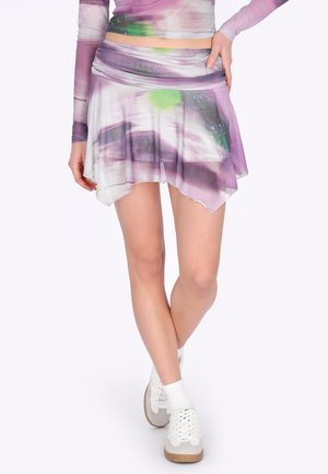 Kobieta w asymetrycznej mini spódnicy z fioletowo-zielonym abstrakcyjnym wzorem, białe trampki oraz dopasowana bluzka z długim rękawem.