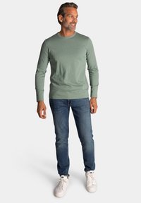 Lichtgroen langslavend shirt, katoenmix, ronde hals, gecombineerd met een blauwe spijkerbroek en witte sneakers, staand tegen een neutrale achtergrond.