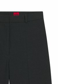 Pantaloni di tessuto scuro con sottili righe verticali, caratterizzati da una vita sagomata e un'etichetta rossa "HUGO" vicino alla parte superiore.